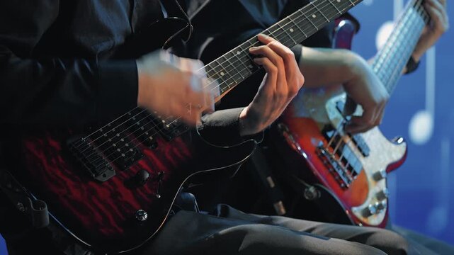A close-up of two guitarists performing on stage. The hands of the musician playing a red and black electric guitar are visible in the foreground. The bass player is in the background