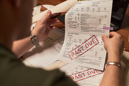 Caucasian young adult man holding utility bills with large past due stamps, sitting at desk reviewing overdue financial documents, visible hands and partial face, focusing on paperwork
