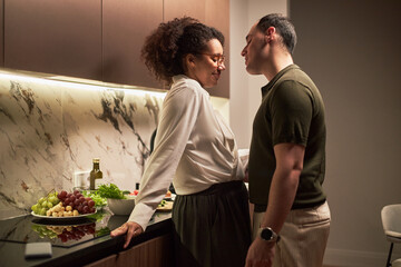Middle aged Black woman and middle aged Caucasian man standing close together in modern kitchen, woman leaning against counter with fresh fruits and vegetables, couple sharing intimate moment