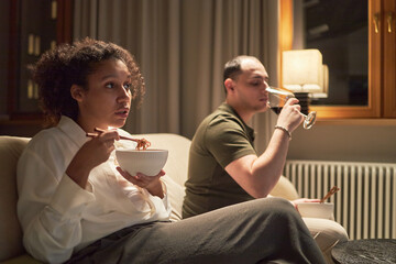 Young adult Black woman eating noodles with chopsticks, sitting on sofa next to young adult Caucasian man drinking from glass, both watching television in living room at night