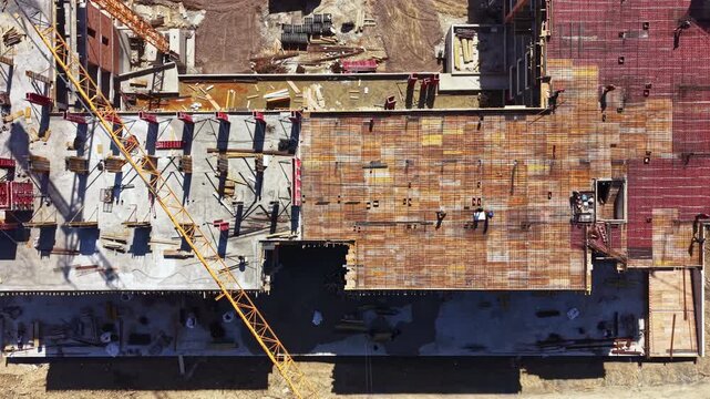 Overview of a construction site with workers placing concrete and structures. Heavy machinery and wooden forms are visible. Building elements and tools are scattered across the area.
