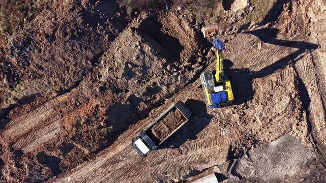A digger is moving dirt at a construction site while a truck waits nearby. The ground shows marks from machinery and broken earth. This scene occurs in daylight hours.