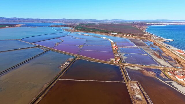 Aerial view shows large salt flats with pools of water in different colors. The coastline is visible in the distance, with land used for salt farming and clear skies above.