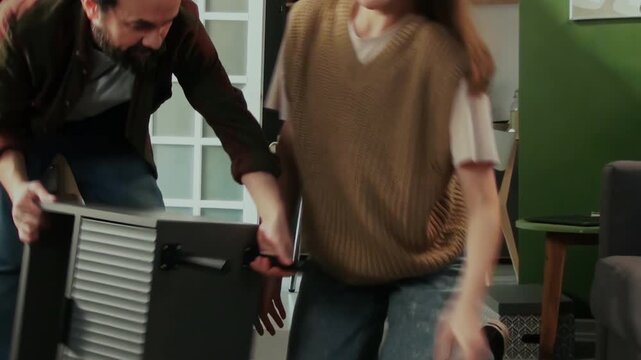 Medium handheld shot of cheerful Caucasian man attaching legs to flatpack drawer, finishing assembly work, turning furniture over, admiring result, hugging and kissing wife with joy and satisfaction