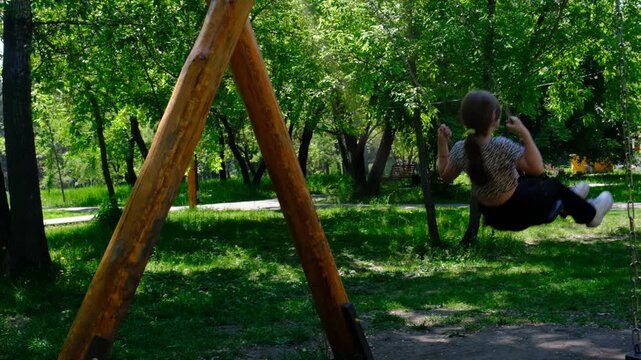 Teenage girl nestled on swing finds solace in the empty park, savours moment of unhurried day. Summer vacations, brightly lit by sunlight and set against a backdrop of lush green trees and sky.