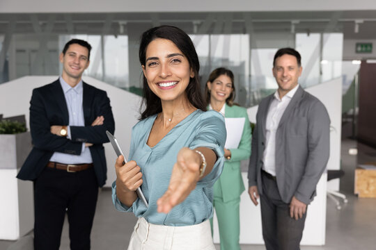 Friendly young woman hr manager recruiter corporate leader standing with diverse business team behind looking at camera with welcoming open palm invite to join company staff start professional career