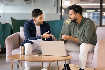Two male colleagues collaborate on task in office lounge, discussing documents or project updates,...