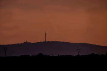 Widok na szczyt Święty Krzyż górę w Górach Świętokrzyskich widok z Ostrowca Świętokrzyskiego. Scena przedstawia zachód słońca, a niebo ma ciepłe, pomarańczowe odcienie. Na szczycie klasztor i wieża tv © Grzegorz