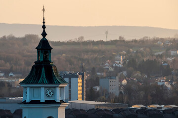Na pierwszym planie wyraźnie widoczna jest wieża kościelna z ciemnozieloną iglicą, na której widnieje zegar. Za nią rozciąga się miejski pejzaż dalej Góry Świętokrzyskie © Grzegorz