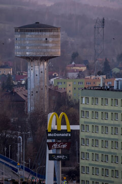 Polska, Ostrowiec Świętokrzyski, 18 marca 2026 17:42. McDonald's &ndash; szyld na betonowym słupie u schyłku zimy na tle wieży ciśnień i budynk&oacute;w mieszkalnych. Szyld przedstawia charakterystyczne ż&oacute;łte łuki