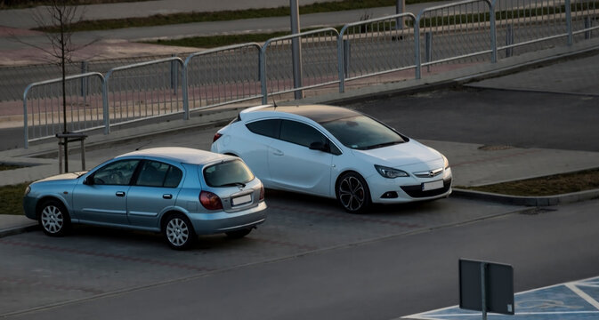 Polska, Ostrowiec Świętokrzyski, 18 marca 2026, 17:32. Samochody osobowe, niebieski Nissan i biały Opel, stoją obok siebie na parkingu. Pojazdy stoją na utwardzonej nawierzchni, obok metalowych barier