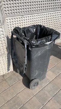 Trash container. Black plastic container on pavement, empty sunlit utility tub lined with black bag against textured wall, shadow creating dramatic contrast, shadows of people. Sunny day