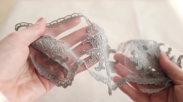 Close-up of hands holding and inspecting a delicate silver-grey embroidered lace trim with a floral damask pattern on a fine mesh background. Perfect for fashion design, and high-end texttile