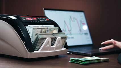 Currency counting machine with stacks of cash and a laptop displaying financial graphs on a wooden...