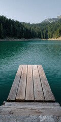 Fototapeta premium Narrow wooden jetty leading into emerald mountain lake surrounded by dense pine forest and distant ridges