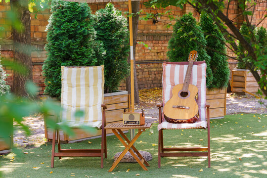 Cozy outdoor relaxation space with wooden garden chairs, acoustic guitar and vintage radio placed on small table. Peaceful backyard leisure scene for music, rest and summer recreation in green garden.