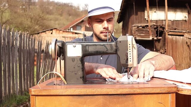 Country style man with hat, suspenders and straw in mouth actively sewing on a vintage machine, carefully guiding fabric while maintaining steady stitching and focused craftsmanship.