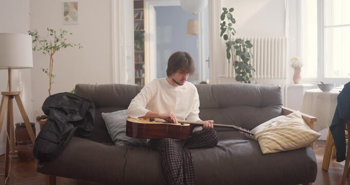 A musician relaxes on a couch, playing guitars and finding unique melodies in a cozy setting