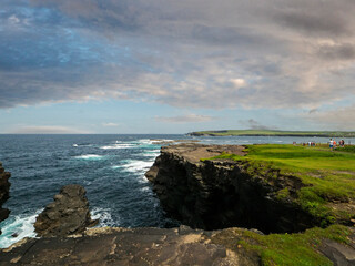 Naklejka premium Rough stone cliff and blue ocean and cloudy sky. Kilkee, county Clare, Ireland. Popular tourist area with amazing Irish nature scenery. Nobody. Warm summer day.