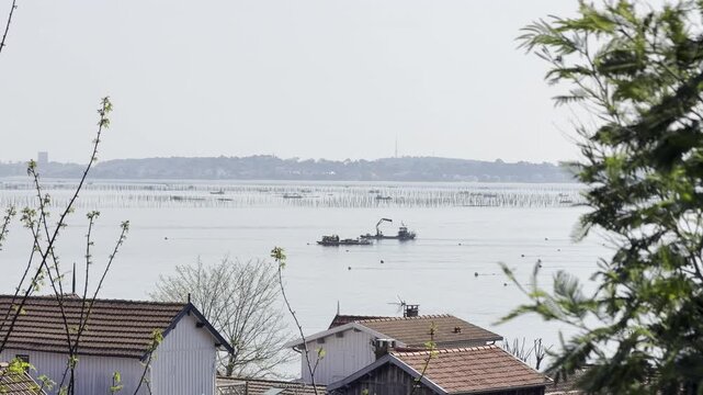 Wide view of Arcachon Basin with an oyster farming boat equipped with a crane in front of oyster beds, with the city of Arcachon in the background, illustrating shellfish industry in southwest France