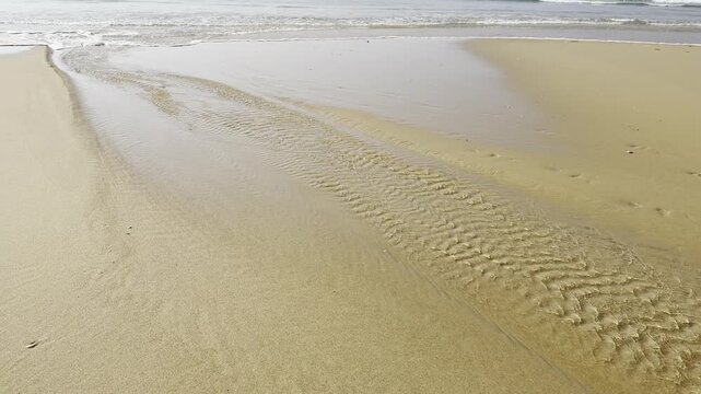 Water flow reversing on the beach as the tide turns, with remaining pools draining toward the ocean meeting incoming waves and shifting back toward the shore