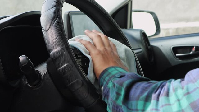 Close up of hands wiping a car steering wheel with a blue cloth, showing interior cleaning, hygiene, and vehicle maintenance routine.
