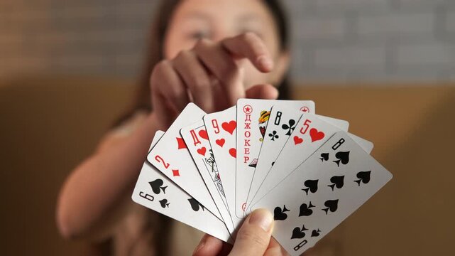 Girl pulls a card from a fan. Close-up view of a hand holding a fan of playing cards, with a young girl in the background choosing the joker card from the deck, concept of gambling, entertainment