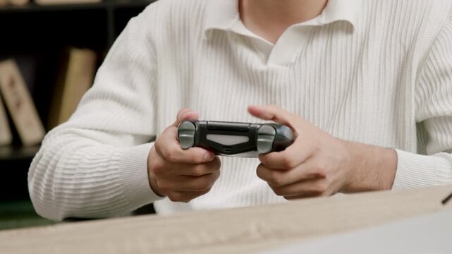 Male gamer in white sweater grips a video game controller with both hands, focused on gameplay, surrounded by bookshelves and a wooden table in a cozy indoor setting