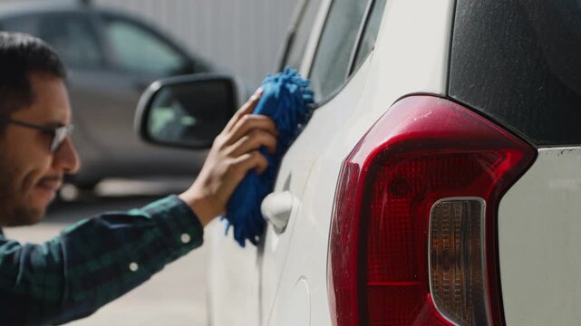 Man wiping the exterior of a car with a blue cloth, showing vehicle cleaning, maintenance, and care routine outdoors.