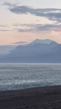 Coastal view of the Mediterranean Sea with the Taurus Mountains in the background. Hazy sunset sky, dark pebble beach in the foreground. Misty mountain peaks.
