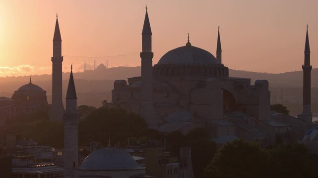The Hagia Sophia mosque with tall minarets stands in silhouette against a soft orange sunrise sky in Istanbul. Gentle morning light creates a calm atmosphere over the historic architecture and