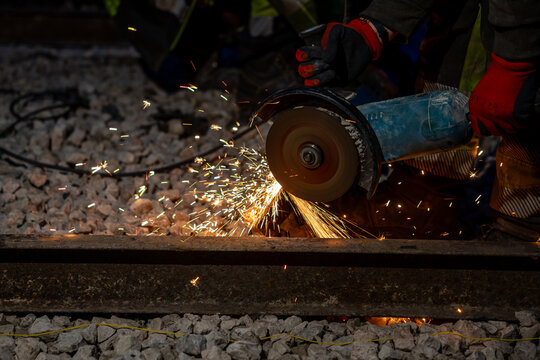 Gloved rail crew uses a handheld abrasive saw to cut a steel rail at night, bright orange sparks spray across crushed ballast, sleepers and rail line are visible.