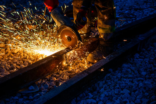 A rail worker in protective gear grinds a steel track, sparks fly over ballast under deep blue twilight. Low directional light and fast shutter emphasize heat and precision.