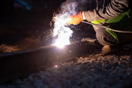 A worker in orange gloves kneels welding a steel rail at night, bright white blue sparks fly, smoke rises, ballast stones lie nearby, low light isolates the weld.