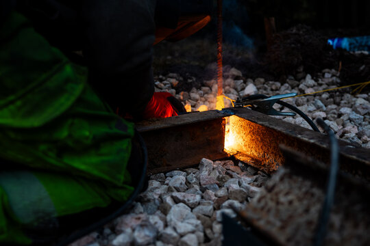 Worker in high visibility gear performs thermite weld on steel rail at night, molten metal glows orange, sparks and smoke rise, small work lamp lights confined trackside scene.