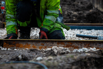 Naklejka premium A worker in a high visibility jacket kneels on mud, aligning a rusted steel edge on crushed stone under overcast light, setting precise levels for concrete or paving.