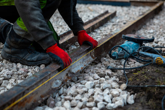 A worker in red gloves kneels on ballast, aligns a rusted rail with a taut string line, measures gauge, and sets a power grinder on damp ground in late day light.