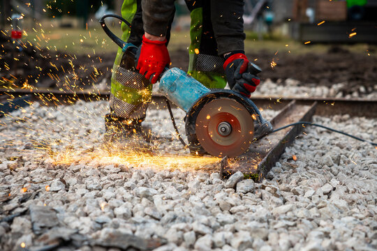 A worker in high visibility pants and red gloves cuts a steel rail with a handheld abrasive saw on ballast, sparks fly, power cable and tools lie nearby in warm low light.