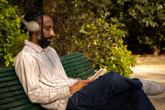 portrait d'un beau jeune homme noir qui lit un livre en &eacute;coutant de la musique avec un casque audio assis sur un banc dans un parc