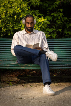 portrait d'un beau jeune homme noir qui lit un livre en &eacute;coutant de la musique avec un casque audio assis sur un banc dans un parc