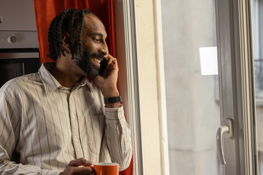 Un homme en t&eacute;l&eacute;travail passe un appel en regardant par la fen&ecirc;tre, caf&eacute; &agrave; la main, dans un moment de r&eacute;flexion calme.