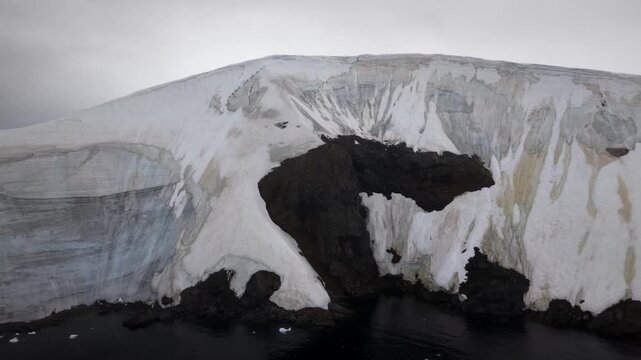 Cinematic aerial raising to reveal a Kelp Gull flying over a prominent basalt rock formation, with the towering, stratified ice cliffs and dark volcanic shores of Elephant Point, Antarctica