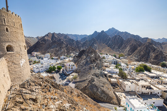Mountain landscape with traditional white houses in Muscat, Oman, viewed from historic fort walls. Dramatic rocky terrain meets urban settlement under clear blue sky, showcasing Middle Eastern archite