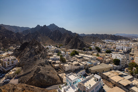 Mountain landscape with traditional white houses in Muscat, Oman, viewed from historic fort walls. Dramatic rocky terrain meets urban settlement under clear blue sky, showcasing Middle Eastern archite