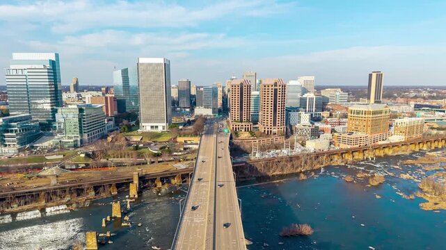 Richmond, Virginia, aerial forward hyperlapse of skyline with cars passing over the Manchester Bridge, James River rapids, and downtown skyscrapers under a clear blue sky.
