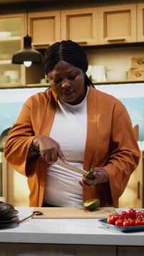 Black content creator, slicing avocado, cooking process, home chef, preparing ingredients on the kitchen counter under natural light. Woman producing high quality food vlog for online channel.