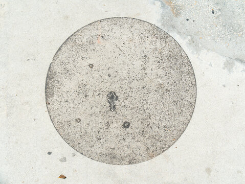 A circular concrete patch or manhole cover is embedded flush within a grey sidewalk pavement showing a grainy porous material.