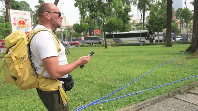 Man With Backpack Exploring City. Individual With Yellow Bag Sightseeing Near Buses In City Park. Traveler In Casual Mood Observing Sights And Reading Route On Mobile Device In Urban Setting
