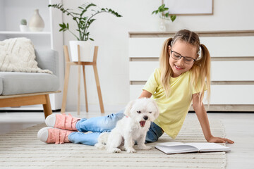 Fototapeta premium Cute little happy girl with Maltese dog reading book on floor at home