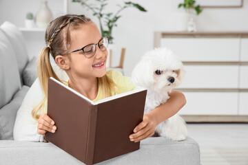 Fototapeta premium Cute little happy girl with Maltese dog reading book on sofa near window at home